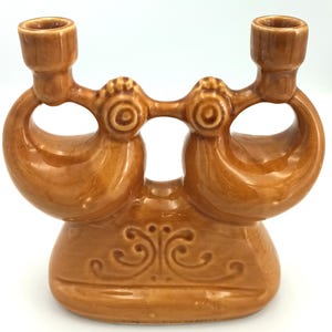 May include: A brown ceramic candle holder with two candle cups. The candle holder has a decorative base with a swirl design. The candle holder has a unique, curved design and is made of glazed ceramic.