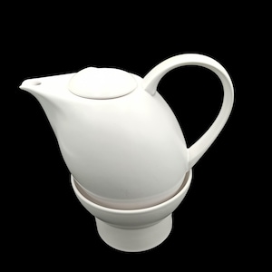Può includere: Una teiera in ceramica bianca con coperchio abbinato e manico curvo. La teiera poggia su una base abbinata, creando un design coerente. Il beccuccio è appuntito e la forma generale è arrotondata.