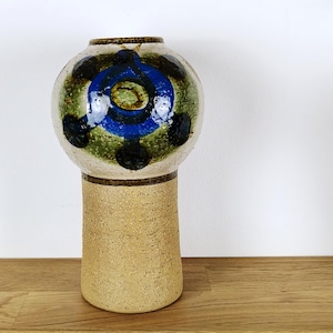 Könnte beinhalten: Eine Keramikvase mit rundem Oberteil und zylindrischem Sockel. Das Oberteil ist mit blauen, grünen und schwarzen kreisförmigen Mustern verziert. Der Sockel hat eine hellbraune Farbe mit strukturierter Oberfläche. Die Vase steht auf einer Holzoberfläche.