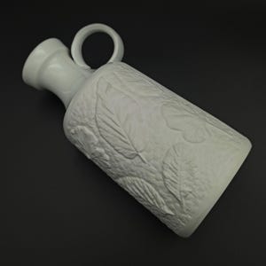 May include: A white ceramic jug with a handle and a textured leaf design. The jug has a long neck and a rounded body. The handle is a simple loop. The design is minimalist and elegant. The jug is suitable for water or other liquids.