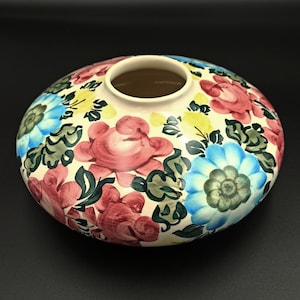 以下が含まれることがあります： 手描きのセラミック花瓶。幅が広く浅い形状です。クリーム色のベースに、赤、青、黄色、緑の色合いの鮮やかな花の模様が施されています。花はフォークアートスタイルで描かれています。