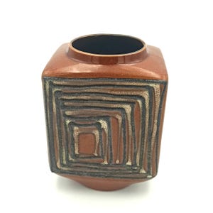 Może przedstawiać: Kwadratowy, ceramiczny wazon w odcieniach brązu z geometrycznym, koncentrycznym kwadratowym wzorem. Wazon ma ciemne wnętrze i zaokrągloną podstawę. Zewnętrzna strona ma teksturowany, warstwowy wzór w kolorze czarnym, beżowym i brązowym.