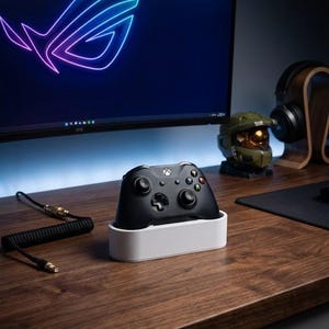 May include: A black wireless game controller rests in a white rectangular charging dock. The controller features a central Xbox button, two analog sticks, a directional pad, and four colored action buttons. A coiled black cable lies nearby.