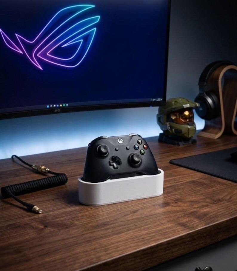 May include: A black wireless game controller rests in a white rectangular charging dock. The controller features a central Xbox button, two analog sticks, a directional pad, and four colored action buttons. A coiled black cable lies nearby.