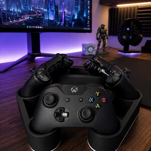 May include: A black plastic charging stand holding three black wireless game controllers. The front controller features a central Xbox logo, a directional pad, two analog sticks, and four colored action buttons labeled A, B, X, and Y.