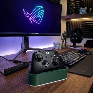 May include: A black wireless game controller rests in a dark green and white rectangular plastic stand. The controller features two analog sticks, a directional pad, and four colored action buttons. It is positioned on a wooden desk surface.