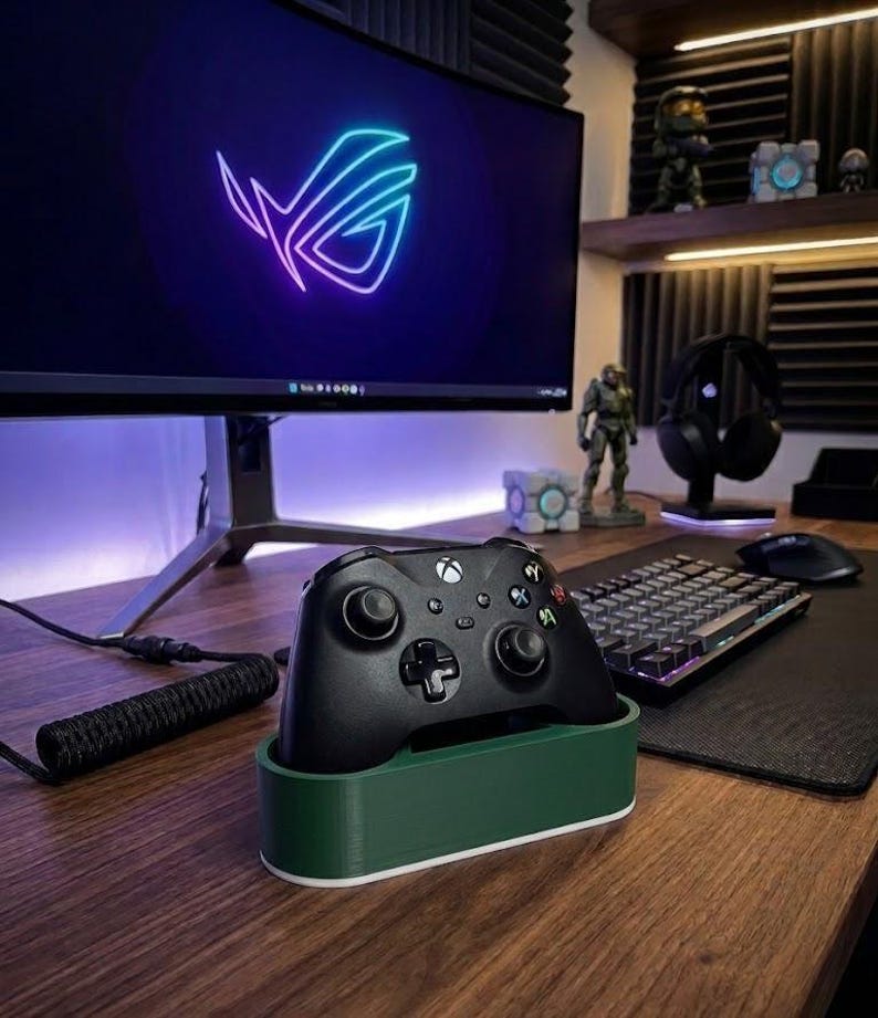 May include: A black wireless game controller rests in a dark green and white rectangular plastic stand. The controller features two analog sticks, a directional pad, and four colored action buttons. It is positioned on a wooden desk surface.