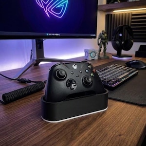 Può includere: Un controller di gioco wireless nero è posizionato su una base di ricarica nera e bianca. Il controller presenta due levette analogiche, un pad direzionale e quattro pulsanti di azione principali etichettati A, B, X e Y su una scrivania in legno.