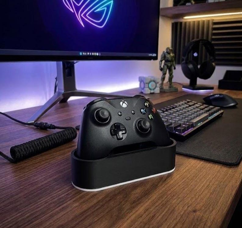 May include: A black wireless game controller rests in a black and white charging dock. The controller features two analog sticks, a directional pad, and four primary action buttons labeled with A, B, X, and Y, positioned on a wooden desk surface.