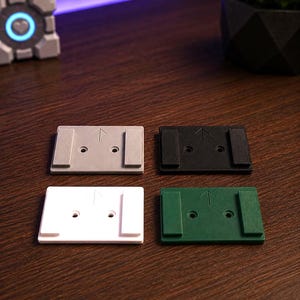 May include: Four rectangular mounting plates in gray, black, white, and green. Each plate features two raised side rails, two central screw holes, and an upward-pointing arrow embossed on the surface. The plates are arranged in a two-by-two grid.
