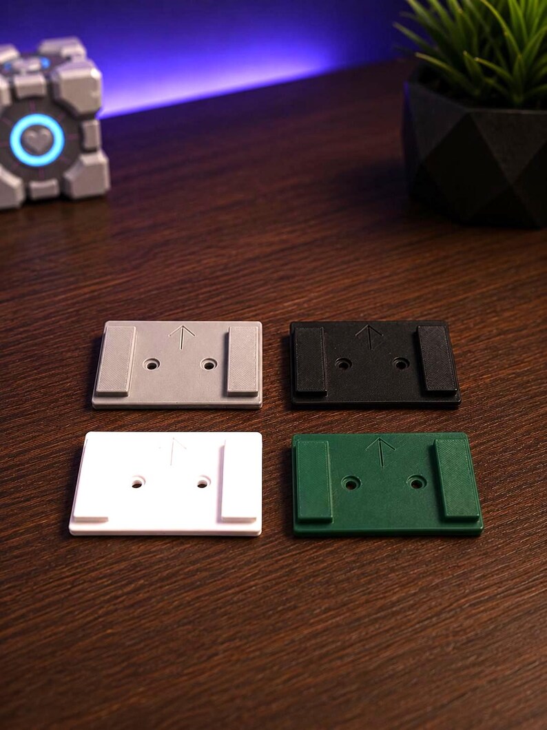 May include: Four rectangular mounting plates in gray, black, white, and green. Each plate features two raised side rails, two central screw holes, and an upward-pointing arrow embossed on the surface. The plates are arranged in a two-by-two grid.