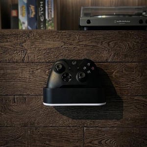 May include: A black wireless video game controller resting in a black charging dock with a white base. The controller features two analog sticks, a directional pad, and four action buttons labeled A, B, X, and Y, positioned on a dark wood-textured surface.