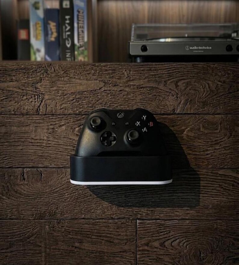 May include: A black wireless video game controller resting in a black charging dock with a white base. The controller features two analog sticks, a directional pad, and four action buttons labeled A, B, X, and Y, positioned on a dark wood-textured surface.