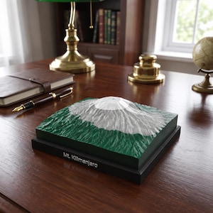 May include: A square 3D topographic model of Mt. Kilimanjaro featuring a white snow-capped peak and textured green slopes. The model is mounted on a black rectangular base with the text Mt. Kilimanjaro printed in white on the front side.