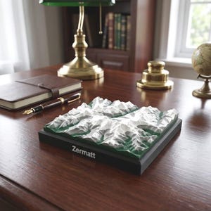 以下が含まれることがあります： ツェルマット山脈の正方形の3D地形レリーフモデル。白い雪に覆われた山頂と緑の谷が特徴です。モデルは黒い台座に取り付けられており、台座の前面の縁には白い文字でZermattと印刷されています。