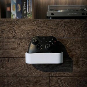 May include: A black wireless video game controller resting inside a rectangular white wall-mounted storage holder. The controller features two analog sticks, a directional pad, and four action buttons labeled A, B, X, and Y on its front surface.