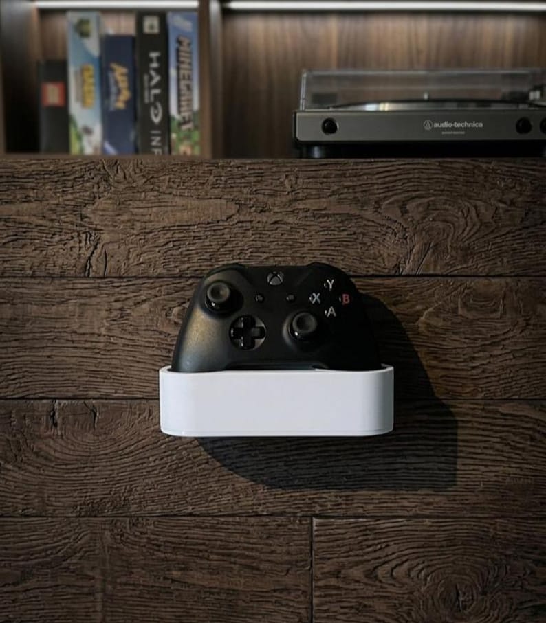 May include: A black wireless video game controller resting inside a rectangular white wall-mounted storage holder. The controller features two analog sticks, a directional pad, and four action buttons labeled A, B, X, and Y on its front surface.