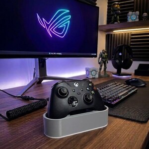 May include: A black wireless game controller rests in a light gray rectangular charging dock. The controller features standard dual analog sticks, a directional pad, and four action buttons. The dock has a smooth, solid-colored exterior finish.