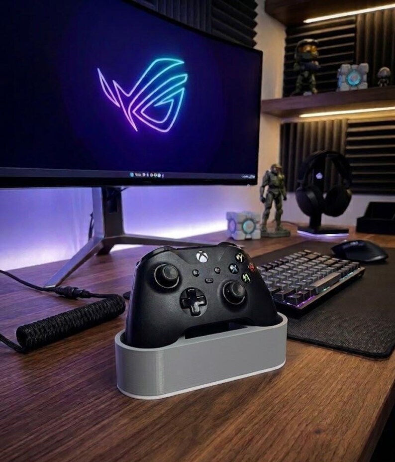 May include: A black wireless game controller rests in a light gray rectangular charging dock. The controller features standard dual analog sticks, a directional pad, and four action buttons. The dock has a smooth, solid-colored exterior finish.