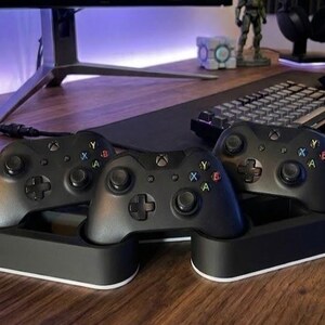 May include: Three black wireless game controllers resting in a dual-sectioned black and white charging dock. Each controller features two analog sticks, a directional pad, and four colored action buttons labeled A, B, X, and Y on the front face.