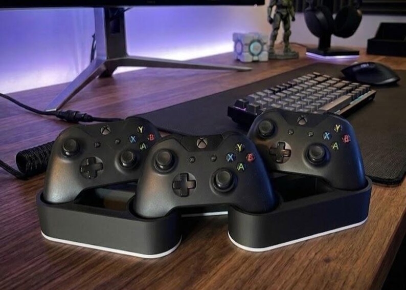 May include: Three black wireless game controllers resting in a dual-sectioned black and white charging dock. Each controller features two analog sticks, a directional pad, and four colored action buttons labeled A, B, X, and Y on the front face.