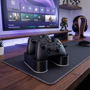 May include: A black dual controller charging stand holding two black wireless game controllers. The stand features a smooth, rounded base with a white accent trim along the bottom edge, designed to securely dock and charge two controllers simultaneously.