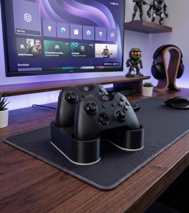 May include: A black dual controller charging stand holding two black wireless game controllers. The stand features a smooth, rounded base with a white accent trim along the bottom edge, designed to securely dock and charge two controllers simultaneously.