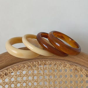 Op de afbeelding: Een set van vier ronde armbanden gerangschikt in een rij. Twee armbanden zijn effen crèmekleurig met een gladde afwerking, terwijl de andere twee een doorschijnend, gemarmerd amber- en bruinpatroon hebben met een glanzend, reflecterend oppervlak.