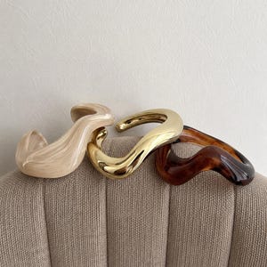 Peut inclure: Trois bracelets ouverts et ondulés disposés côte à côte. Le premier est en résine marbrée beige, le deuxième en métal doré poli et le troisième en résine à motif écaille de tortue brun foncé. Chaque pièce présente une forme fluide et sinueuse.