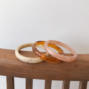 Pode incluir: Três pulseiras circulares com um acabamento liso e polido. Uma pulseira é de cor creme sólida, uma é âmbar translúcida com padrões castanhos marmoreados e uma é rosa pálido com uma aparência subtil, nublada e semitransparente.