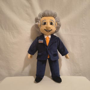 Può includere: Una bambola di peluche che raffigura un personaggio con capelli grigi, che indossa un abito blu scuro, una camicia bianca con colletto e una cravatta arancione. La bambola ha una piccola toppa rettangolare sul petto con le lettere TIG in blu.