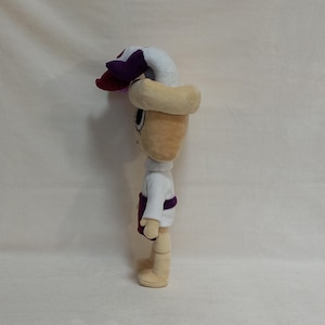 May include: A plush doll with a beige head and limbs, wearing a white shirt, purple sash, and a beige hat with a white and purple flower. The doll has large eyes and a smiling expression. The doll is standing upright.
