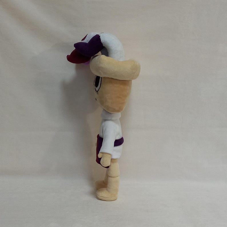 May include: A plush doll with a beige head and limbs, wearing a white shirt, purple sash, and a beige hat with a white and purple flower. The doll has large eyes and a smiling expression. The doll is standing upright.
