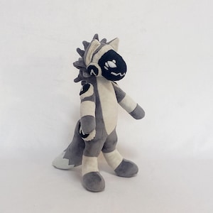 May include: A plush toy featuring a grey and cream-colored creature with a dark visor-like face mask. The character has a spiky mane, a long tail, and circular patches on the shoulders and thighs. The design includes soft fabric textures throughout.