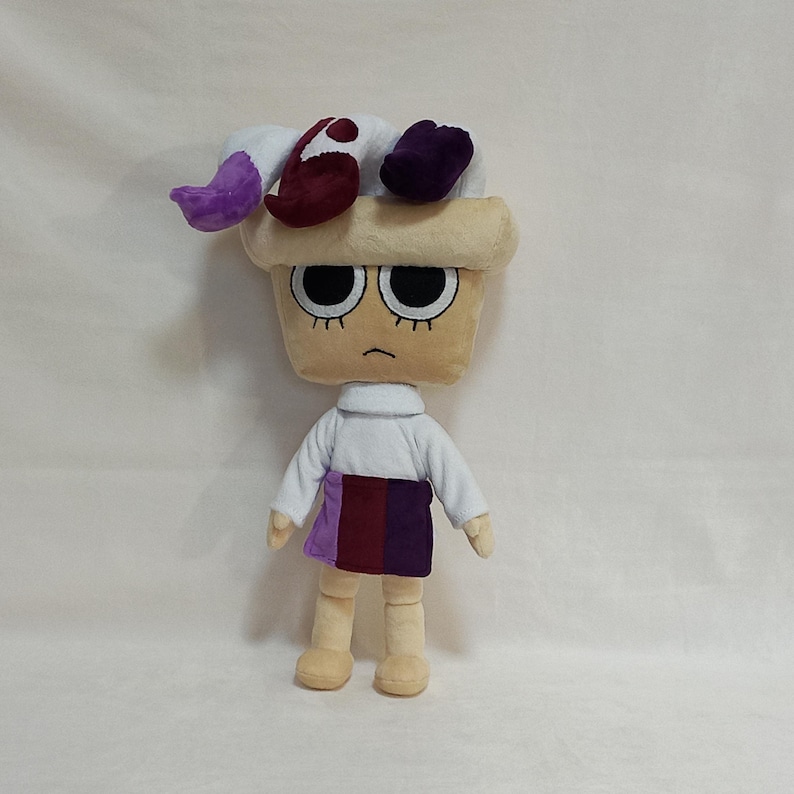 May include: A plush doll with a tan, cupcake-shaped head, large black eyes, and a grumpy expression. It wears a white shirt, a purple and burgundy skirt, and a whimsical hat with purple, white, and burgundy accents. The doll has tan arms and legs.