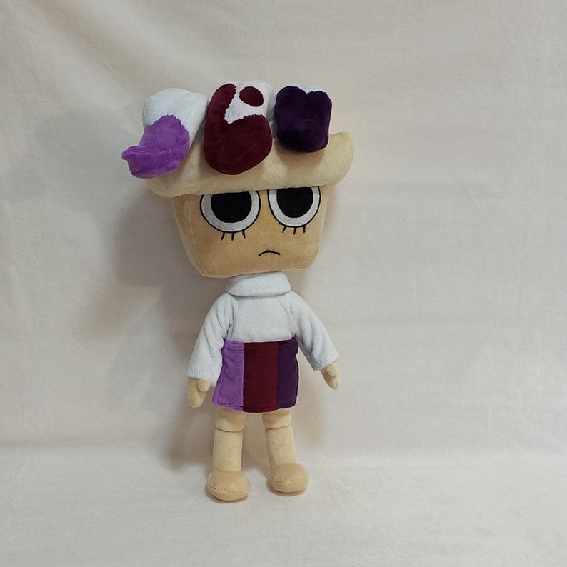 May include: A plush doll with a cupcake-shaped head, large black eyes, and a slightly frowning mouth. The doll wears a white long-sleeved shirt, a purple and burgundy skirt, and beige legs. The head has a cream-colored base with purple, burgundy, and white accents.