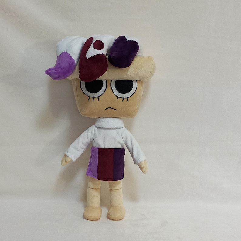May include: A plush doll with a tan, square-shaped head, large black eyes, and a neutral expression. The doll has a white turtleneck shirt, a purple and burgundy skirt, and tan legs and arms. The doll's hair is a mix of white, purple, and burgundy.