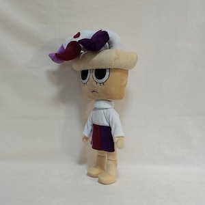 May include: A plush doll with a tan head and body, wearing a white turtleneck and a skirt with red and purple stripes. The doll has black eyes and a sad expression. A white and purple floral-shaped accessory sits atop the head.