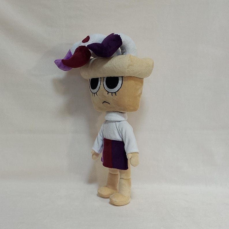 May include: A plush doll with a tan head and body, wearing a white turtleneck and a skirt with red and purple stripes. The doll has black eyes and a sad expression. A white and purple floral-shaped accessory sits atop the head.