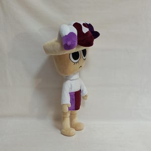 May include: A plush doll with a beige head and body, wearing a white turtleneck, a purple and maroon skirt, and beige shoes. The doll has a large, cream-colored hat with white, purple, and maroon accents. The doll has large, black eyes and a neutral expression.