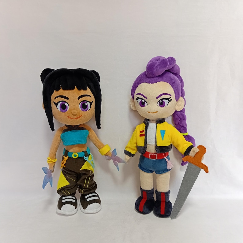 May include: Two plush dolls with cartoon-like features. One doll has black hair, a turquoise top, and brown pants. The other doll has purple hair, a yellow jacket, and blue shorts. Each doll holds a weapon.