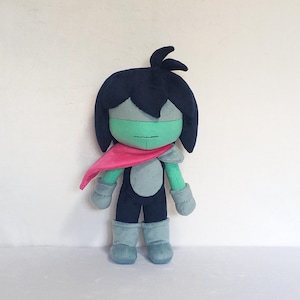 May include: A plush doll of a character with a dark blue head and hair, a light green face, and a pink scarf. The doll is wearing a dark blue outfit with gray accents and light blue boots and gloves. The doll is standing upright.