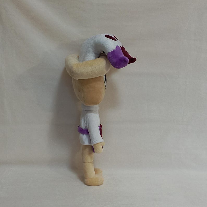 May include: A plush doll with a light tan head and body, wearing a white shirt with a purple sash. The doll has a light tan hat and a white and purple headpiece. The doll is standing upright.