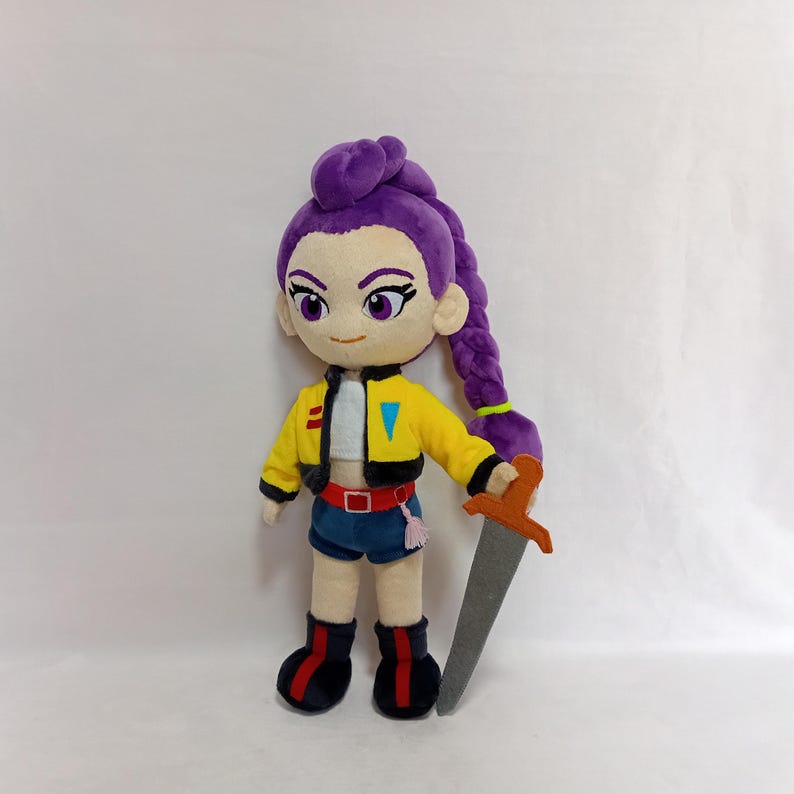 May include: A plush doll of a character with purple hair in a long braid, wearing a yellow jacket, blue shorts, and black boots with red accents. The doll is holding a felt sword with an orange handle.