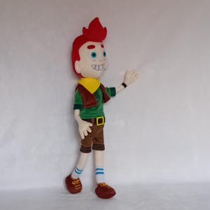 May include: A plush doll of a cartoon character with red hair, a yellow neckerchief, and a green shirt. The doll is wearing brown shorts, brown shoes, and blue and white striped socks. The doll is waving with its right hand.