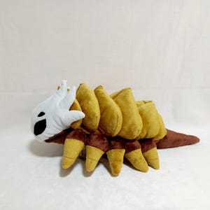 May include: A plush toy in the shape of a stylized insect. The toy features a white head with black eye details, a segmented mustard-yellow body, and brown legs and tail. The plush is soft and appears to be made of fabric.