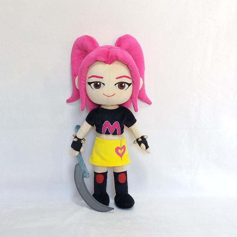 May include: A plush doll with pink hair styled in two high ponytails. The doll wears a black crop top with a pink "M" and a yellow skirt with a heart. It holds a gray scythe and has black wristbands.