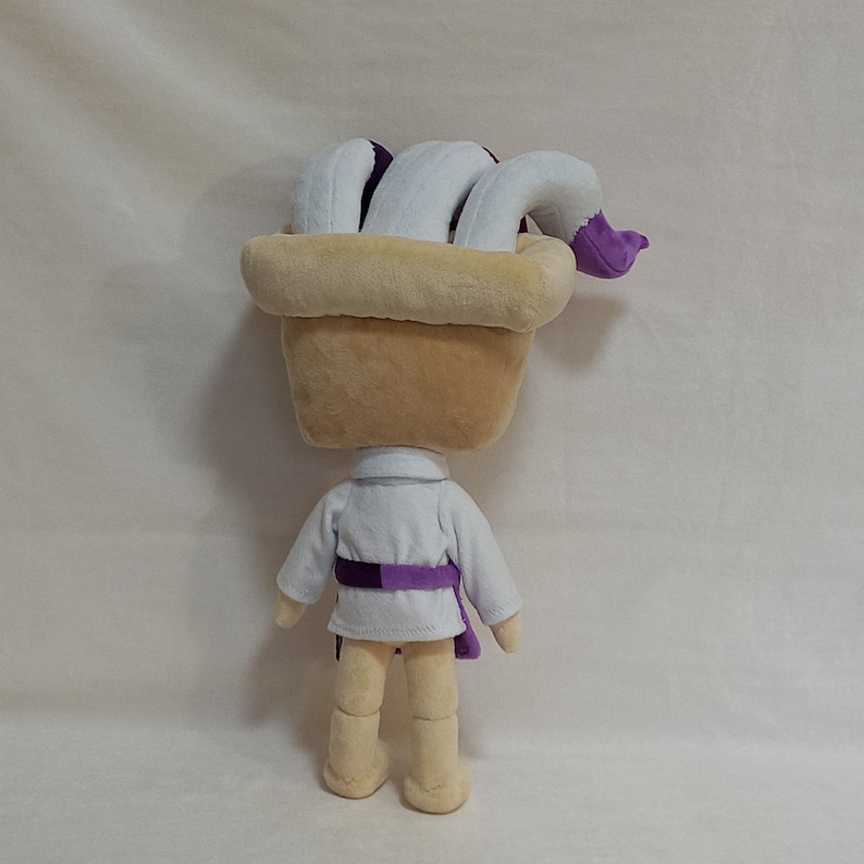 May include: A plush doll with a tan head and body, wearing a white shirt with a purple belt. The doll has a large, tan hat with white and purple accents. The doll's legs are tan.