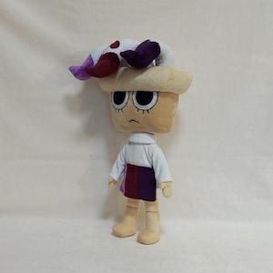 May include: A plush toy character with a tan, square-shaped head and large, black eyes. It wears a white shirt, a skirt with purple and burgundy panels, and a headpiece with purple, white, and burgundy accents. The toy has a sad expression.