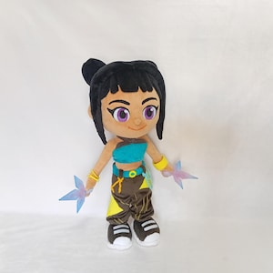 May include: A plush doll of a character with black hair, purple eyes, and a teal top. The doll wears brown pants with yellow accents and white shoes. It holds two blue and pink star-shaped objects.